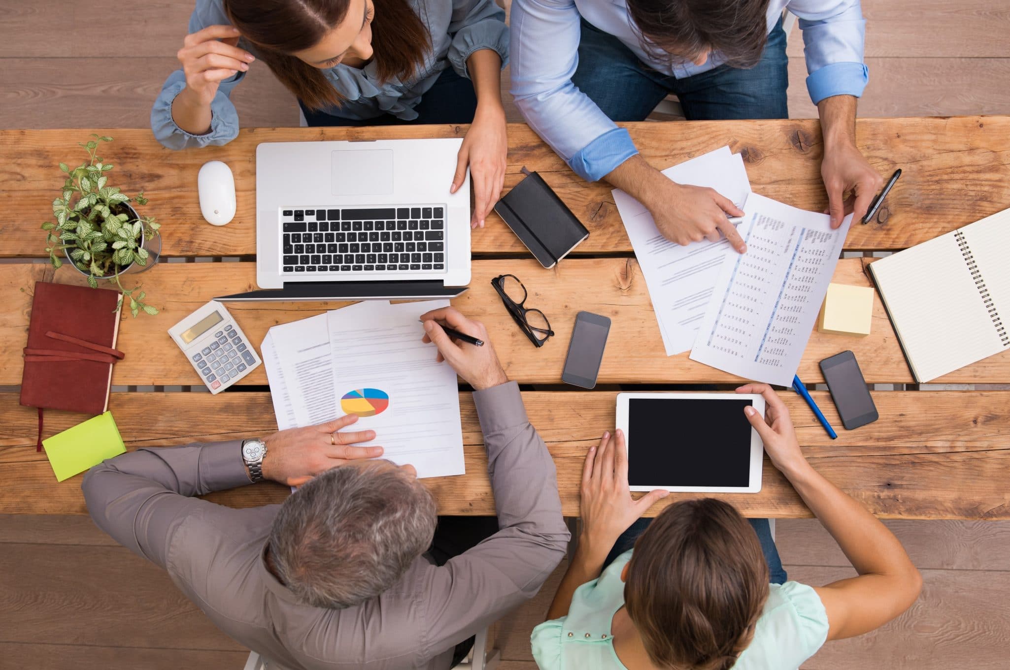  A group of people are sitting around a table and working together on a project. Some of them are looking at a laptop, while others are looking at papers. One person is writing something down on a notepad.